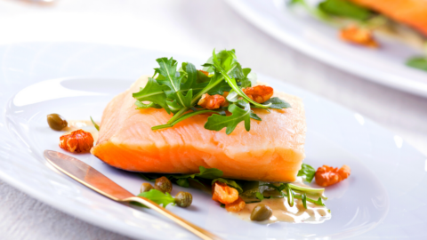 A piece of cooked salmon topped with arugula, nuts, and capers is served on a white plate with a fork and knife.