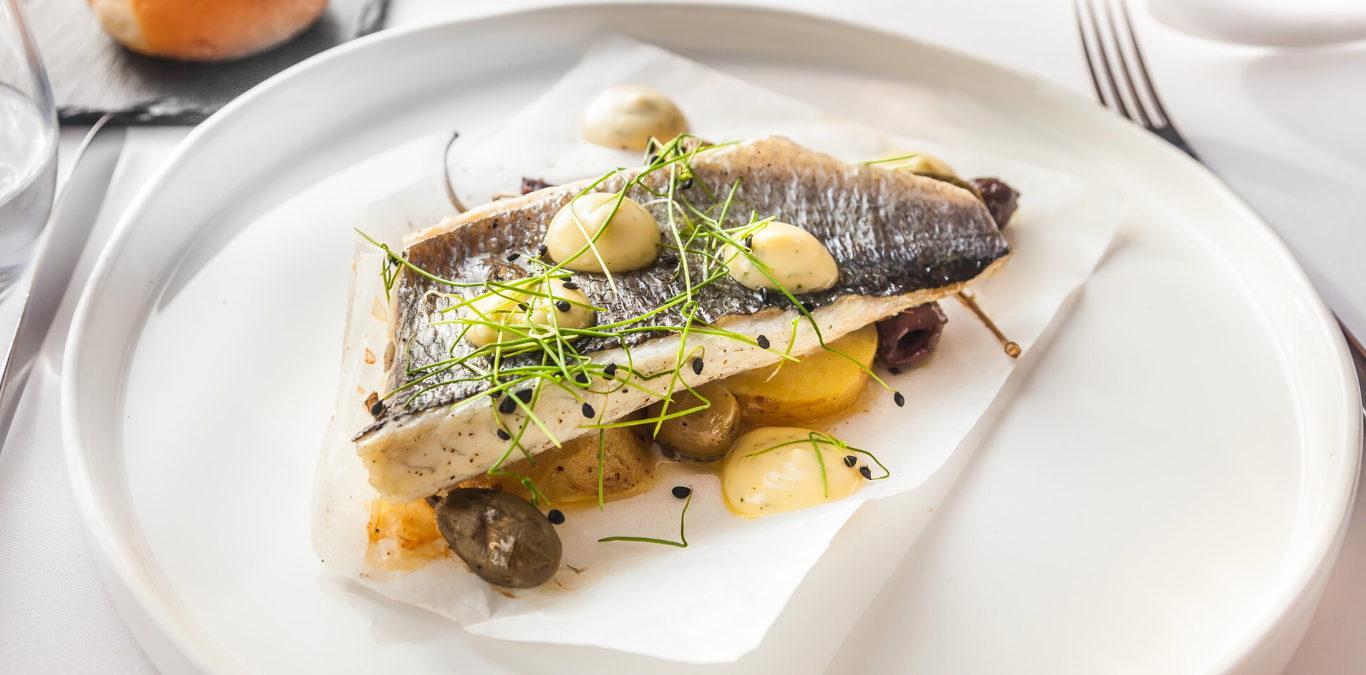 A plated fish fillet served on top of sliced potatoes, garnished with herbs, olives, and dollops of creamy sauce.