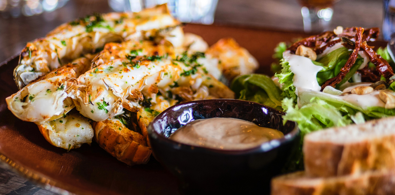 A plate with grilled lobster tails, salad with dressing, sliced bread, and a small bowl of dipping sauce.