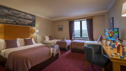 Hotel room with one double bed and two single beds, a window with a scenic view, TV displaying a cartoon, a desk with toys, and warm-toned decor.