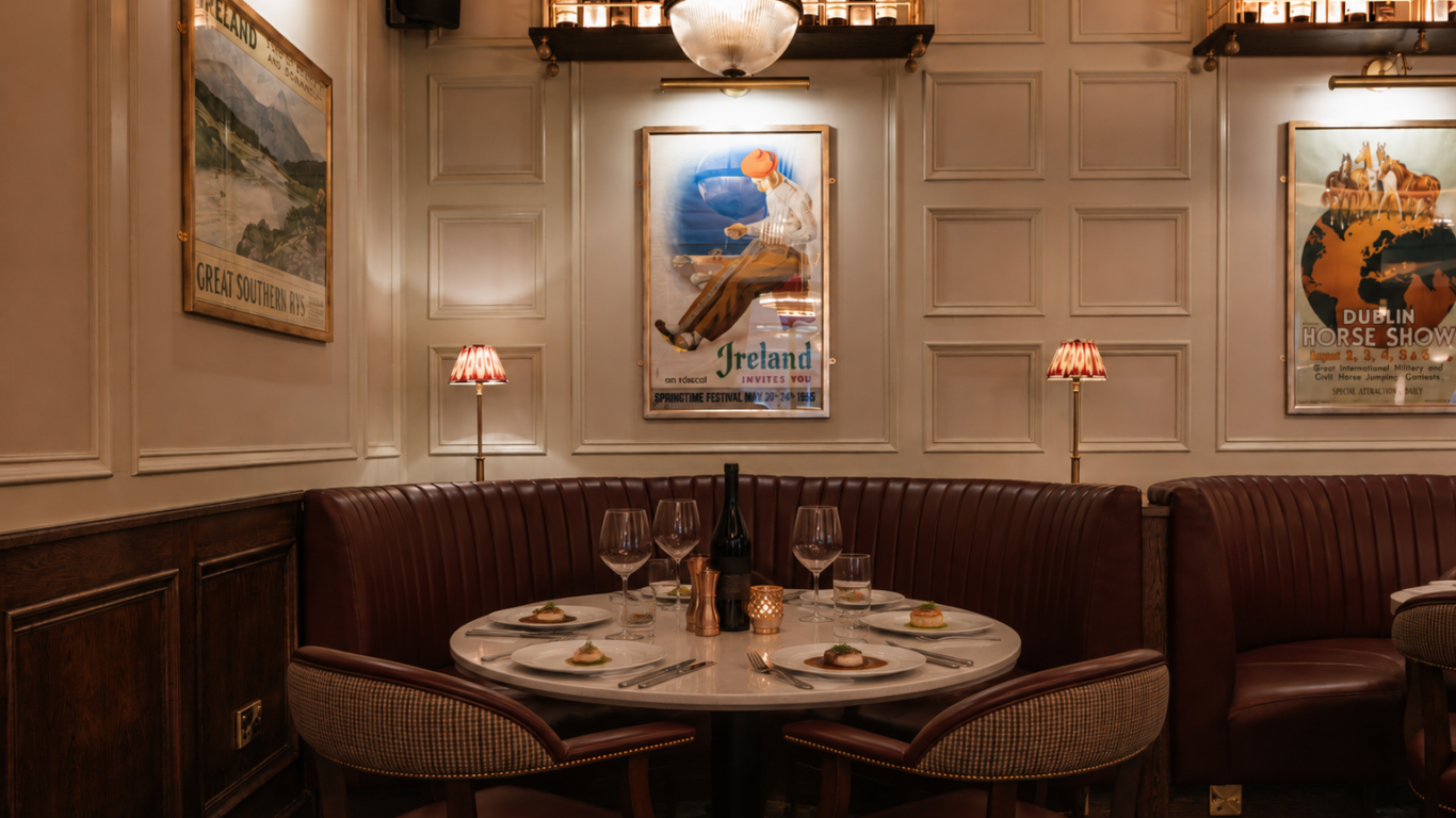 A dimly lit restaurant booth with a round table set for four, wine glasses, plates, a candle, and vintage travel posters on the wall.