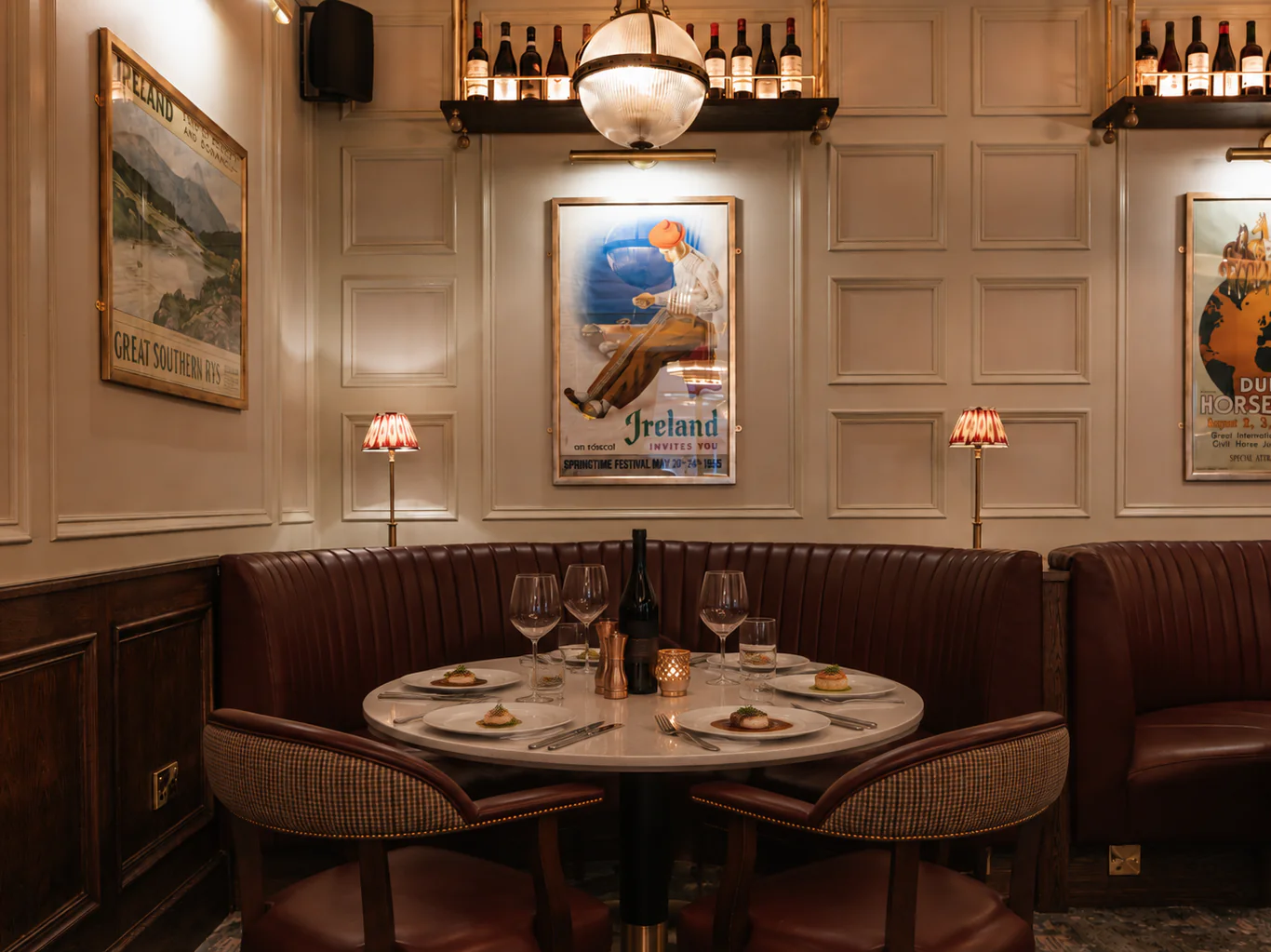 A dimly lit restaurant booth with a round table set for four, wine glasses, plates, a candle, and vintage travel posters on the wall.