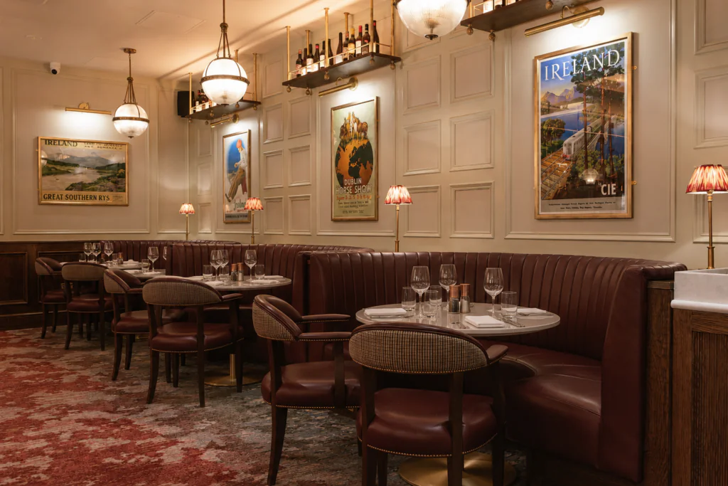 Carriage 3 Elegant restaurant interior with round tables, red leather booths, vintage travel posters on cream paneled walls, and soft lighting from hanging globes and small table lamps.
