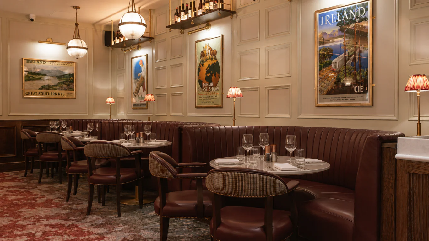 Elegant restaurant interior with round tables, red leather booths, vintage travel posters on cream paneled walls, and soft lighting from hanging globes and small table lamps.