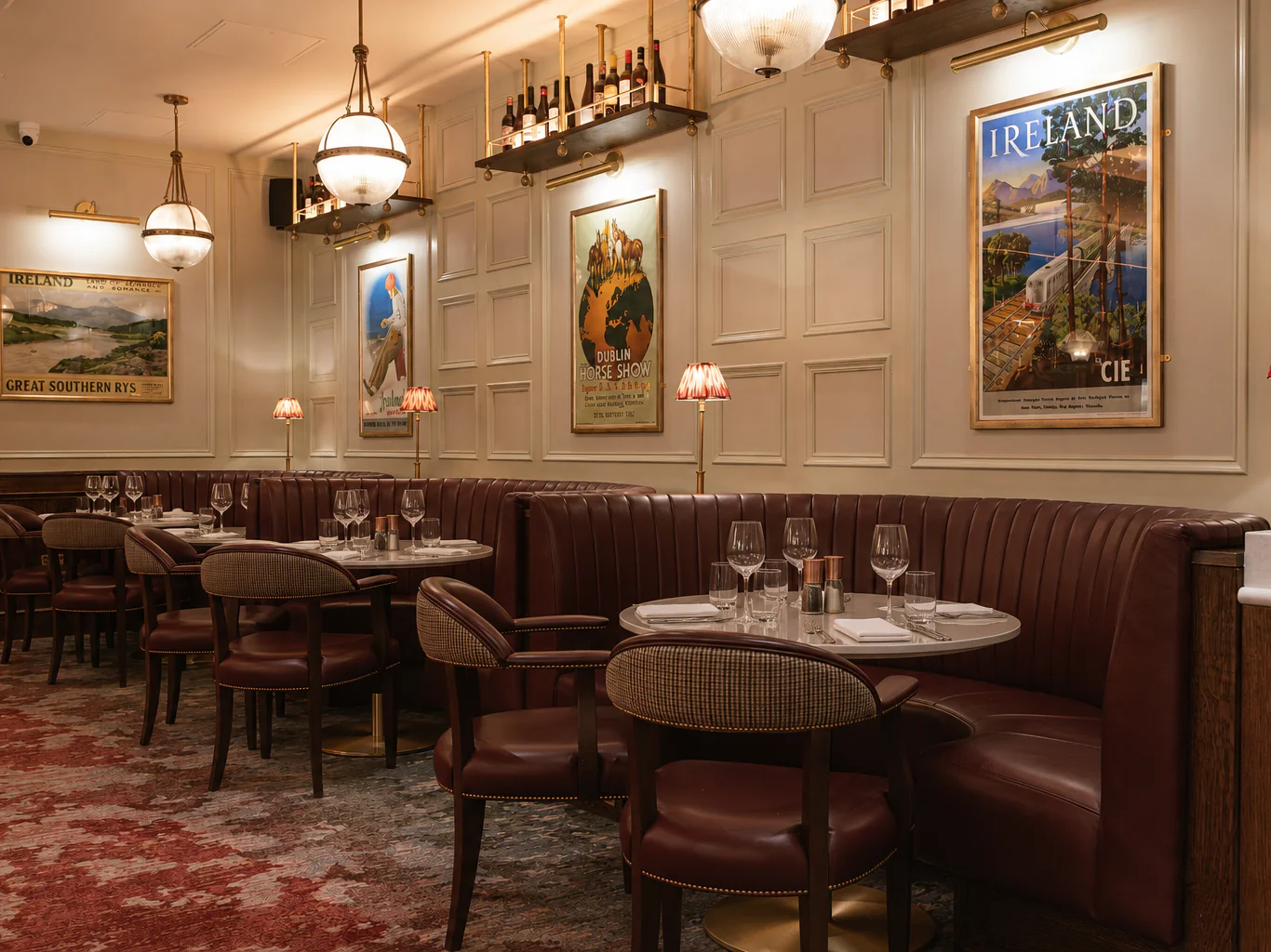 Elegant restaurant interior with round tables, red leather booths, vintage travel posters on cream paneled walls, and soft lighting from hanging globes and small table lamps.