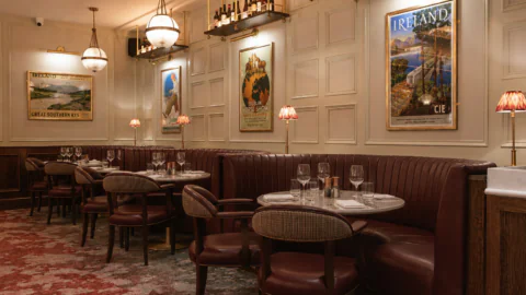 Elegant restaurant interior with round tables, red leather booths, vintage travel posters on cream paneled walls, and soft lighting from hanging globes and small table lamps.