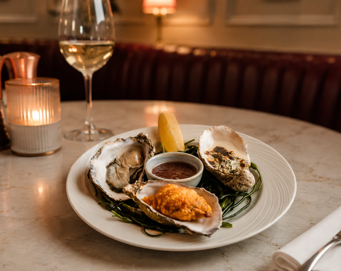A plate with three oysters, a lemon wedge, sauce, and a glass of white wine on a restaurant table with a lit candle.