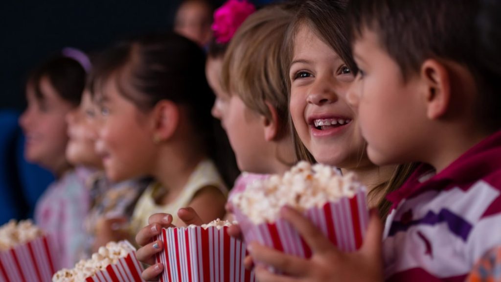 A group of children sit in a row, smiling and holding striped popcorn containers, watching something off-screen.