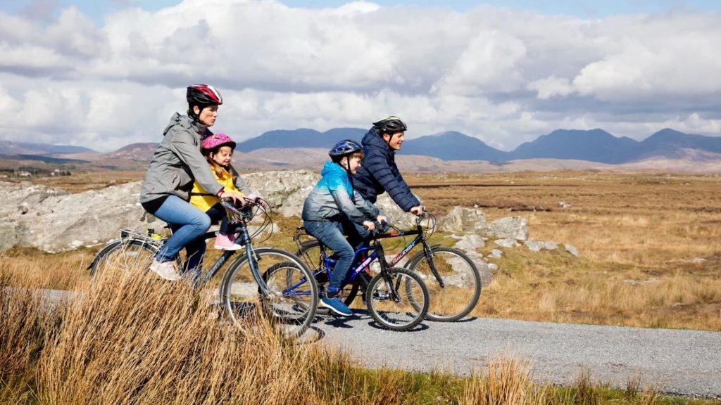 A family of four, wearing helmets and jackets, ride bicycles on a paved path through a grassy landscape with mountains in the background.