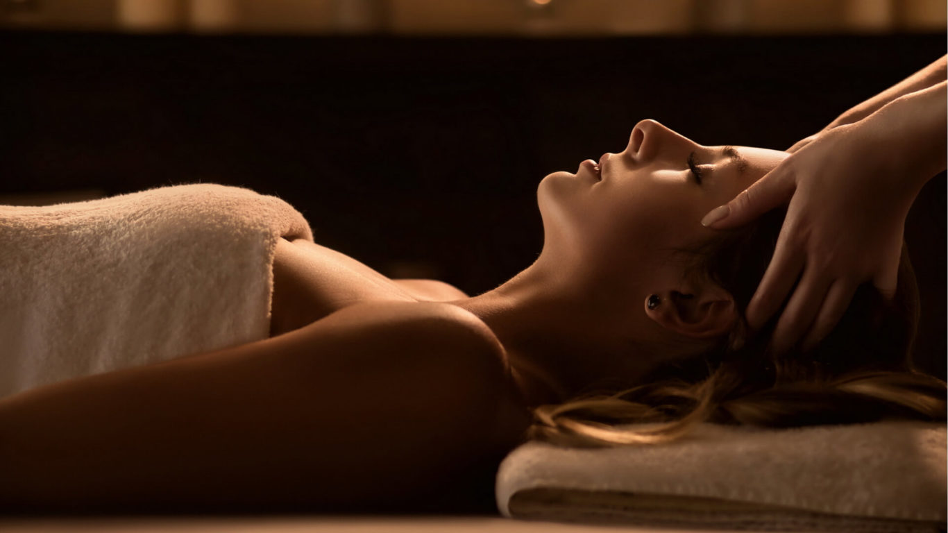 A person lies on a massage bed with eyes closed, covered by a towel, while receiving a head massage from another individual.