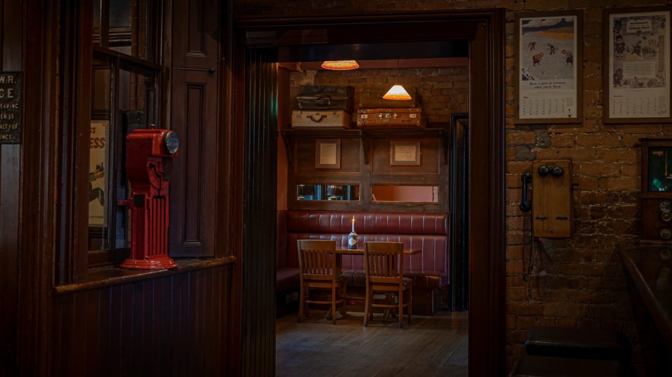 A dimly lit vintage pub interior with a wooden table, four chairs, red upholstered booth seating, brick walls, and old suitcases on a shelf.
