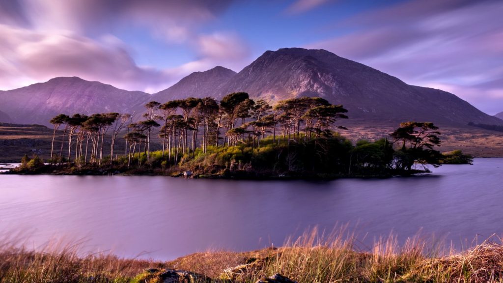 A small tree-covered island sits in a calm lake with mountains in the background under a partly cloudy sky at dusk.