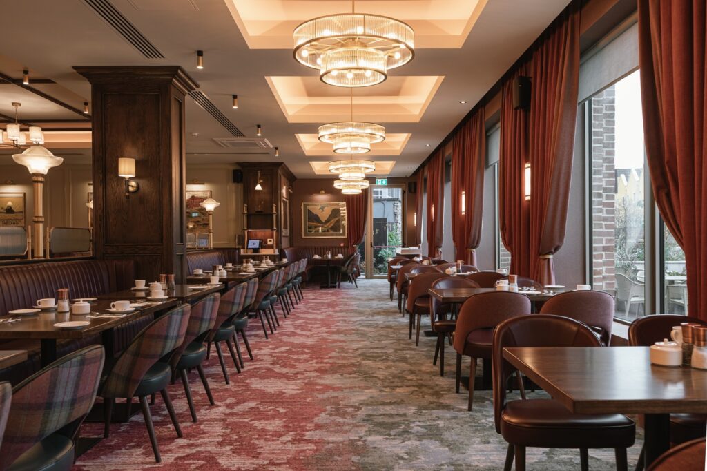 Elegant restaurant interior with rows of tables and chairs, red carpet, large windows with curtains, and modern ceiling lights. Tables are set with cups and saucers.