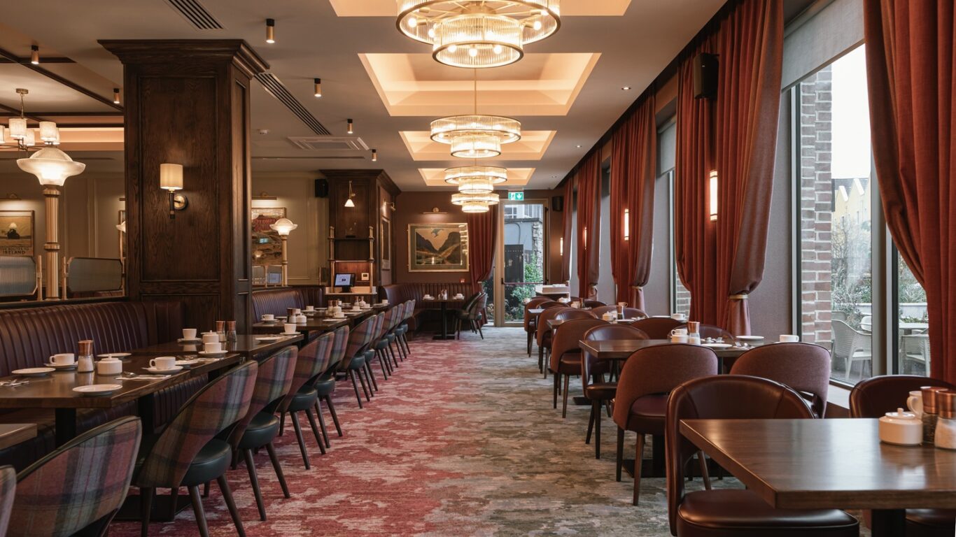 Elegant restaurant interior with rows of tables and chairs, red carpet, large windows with curtains, and modern ceiling lights. Tables are set with cups and saucers.