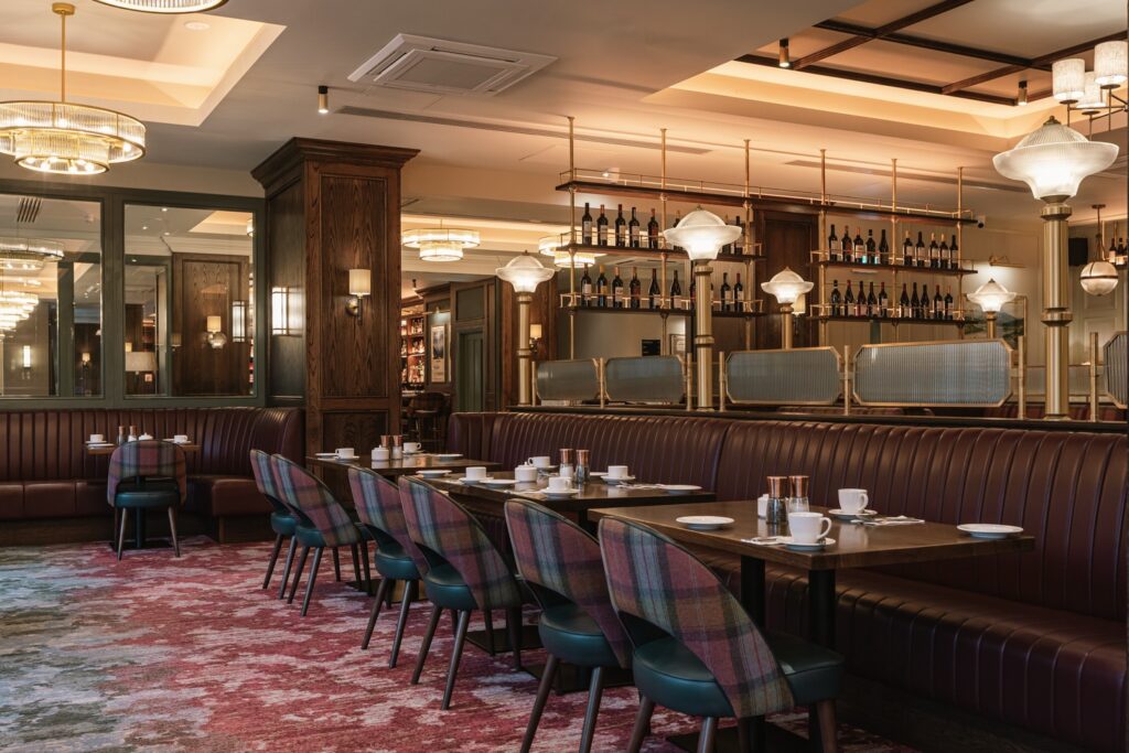 A modern restaurant interior with brown leather banquettes, patterned chairs, set tables, carpeted flooring, and shelves stocked with wine bottles behind glass dividers.