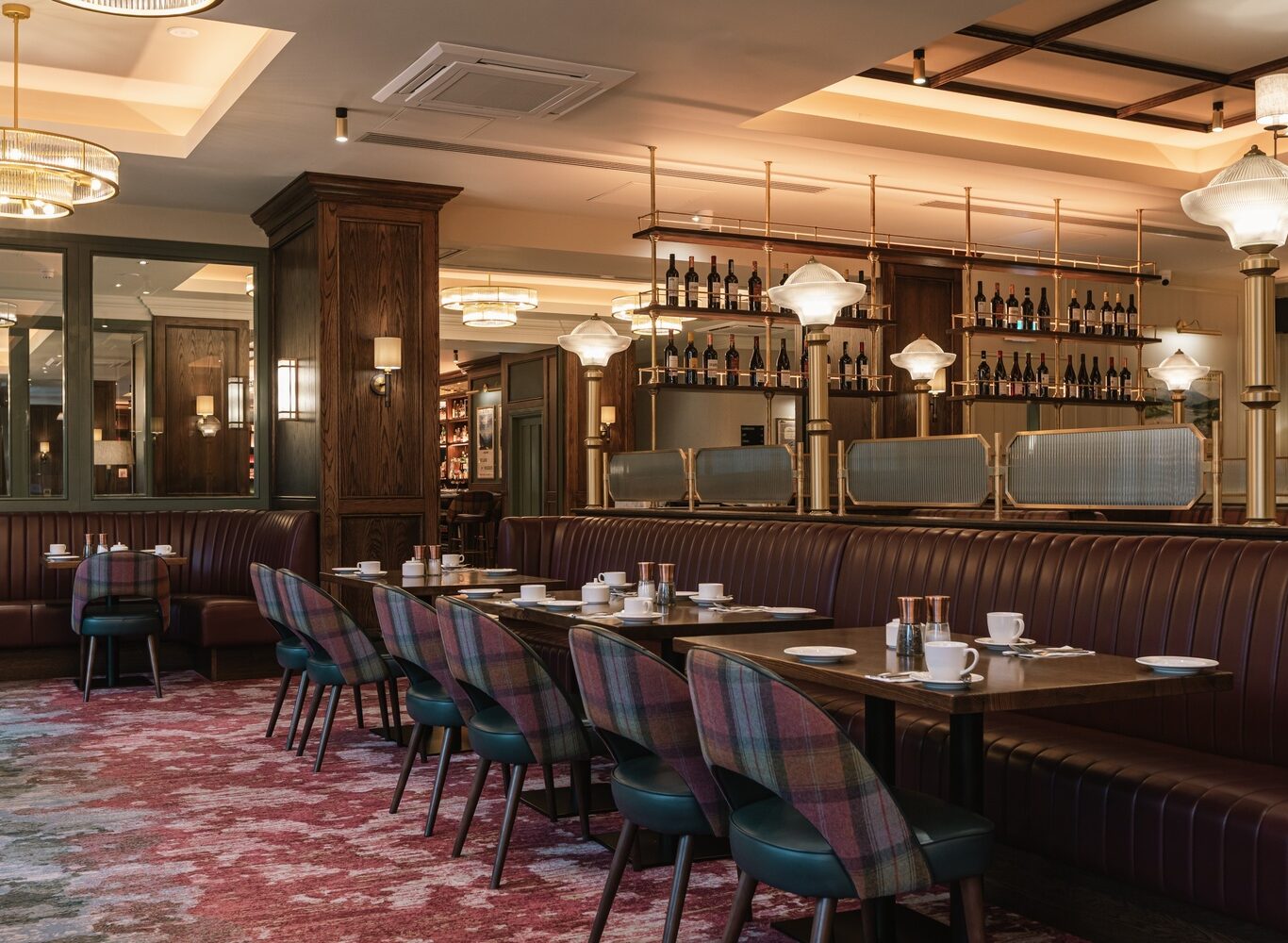 A modern restaurant interior with brown leather banquettes, patterned chairs, set tables, carpeted flooring, and shelves stocked with wine bottles behind glass dividers.