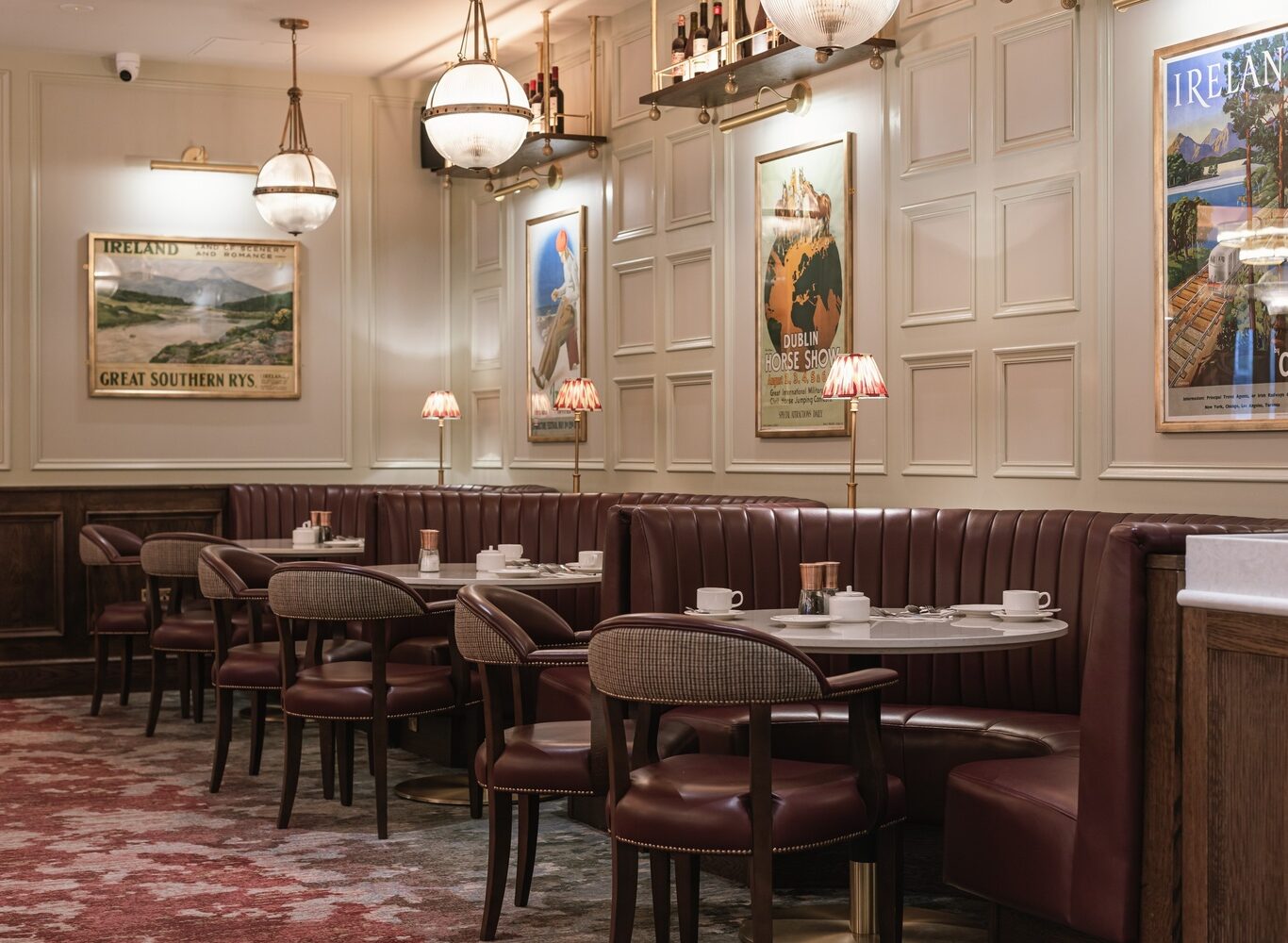 A cozy restaurant with maroon booths, round tables set with cups and saucers, wall art, vintage lamps, and paneled walls.
