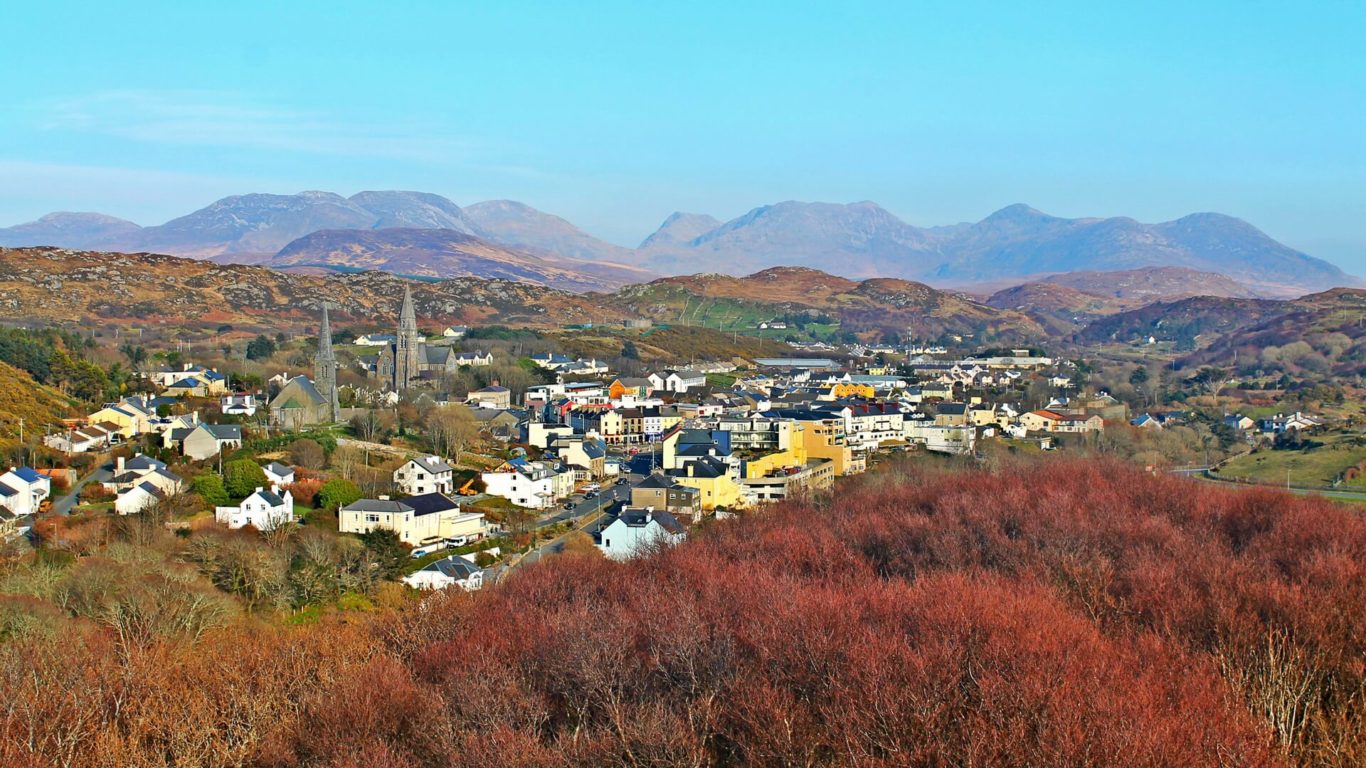 A small town with white and yellow buildings is nestled among hills with reddish-brown and green vegetation, with mountains visible in the distance under a blue sky.