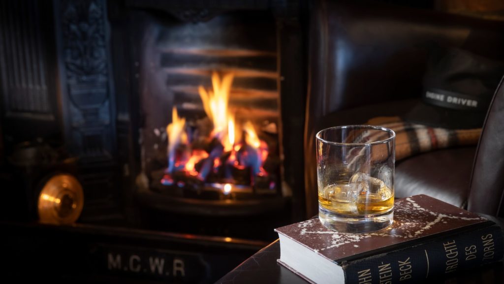 A glass of whiskey with ice sits on two books in front of a lit fireplace, with a hat resting on a nearby leather chair.