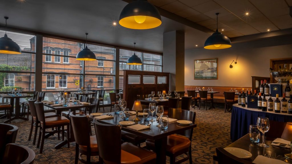 A warmly lit restaurant dining area with set tables, wooden chairs, large windows, and a wine display table in the foreground.
