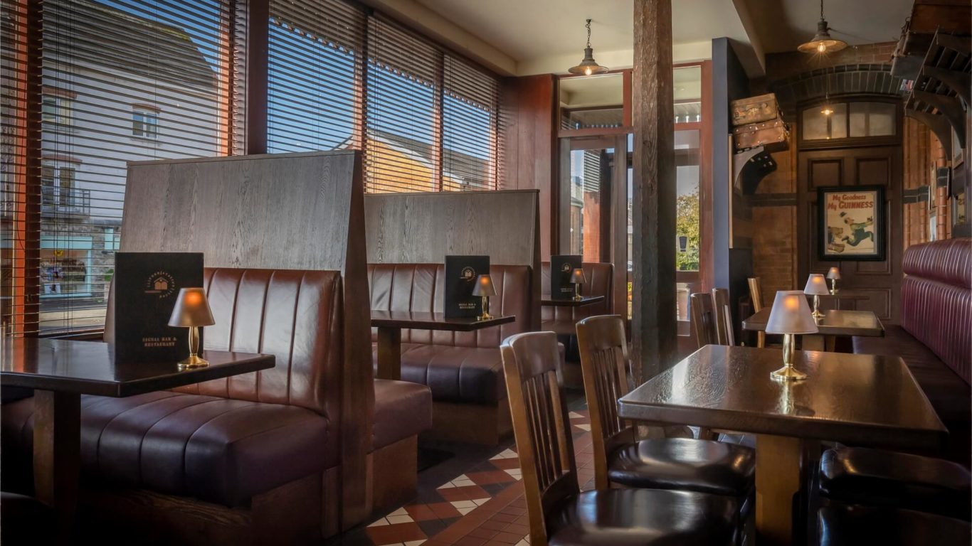Interior of a restaurant with wooden booths, tables, chairs, table lamps, and large windows with blinds letting in natural light.