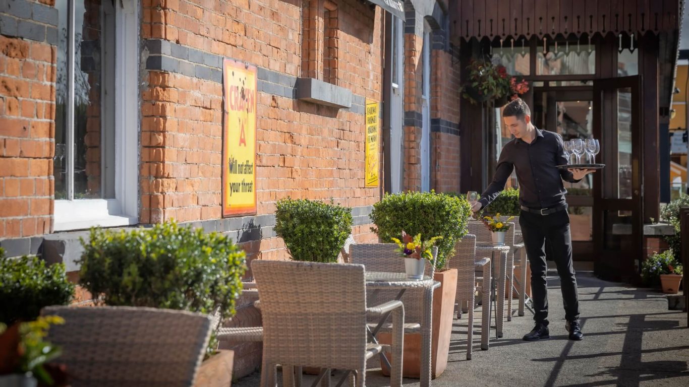 A waiter carries a stack of glasses while setting up an outdoor seating area with wicker chairs and potted plants against a brick building.