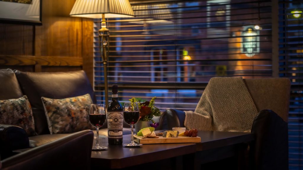 A cozy living room scene with a table set for two, featuring glasses of red wine, a bottle, cheese, and charcuterie, beside a sofa and armchair under warm lighting.