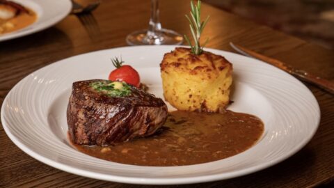 A plate with a grilled steak topped with herb butter, a serving of potato gratin, brown sauce, and a roasted cherry tomato, set on a wooden table.