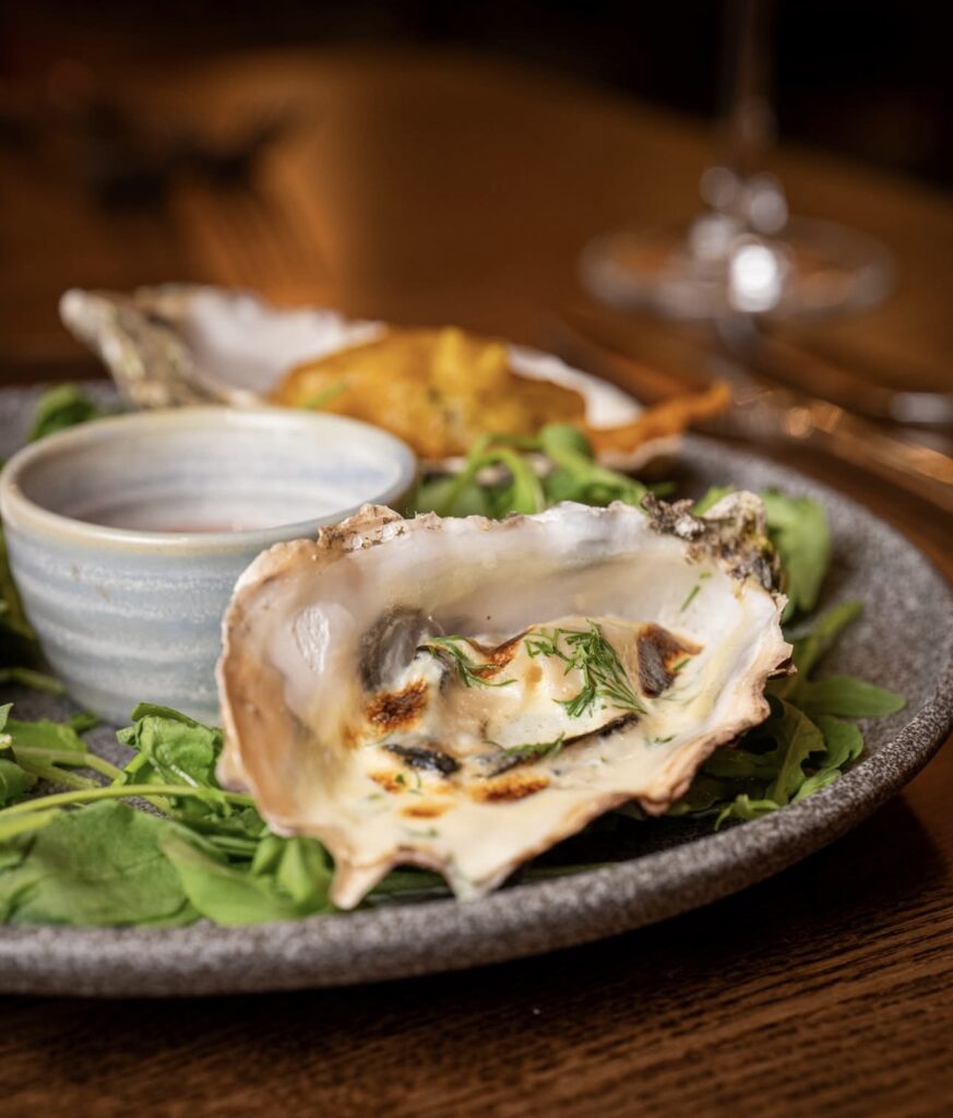 A grilled oyster with herbs is served on a bed of greens alongside a small cup of sauce on a gray plate.