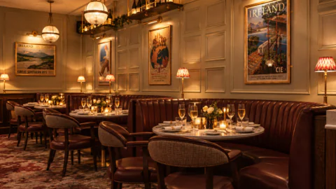 A dining area with round tables, dark leather booths, and chairs, decorated with framed vintage Irish travel posters on the walls.