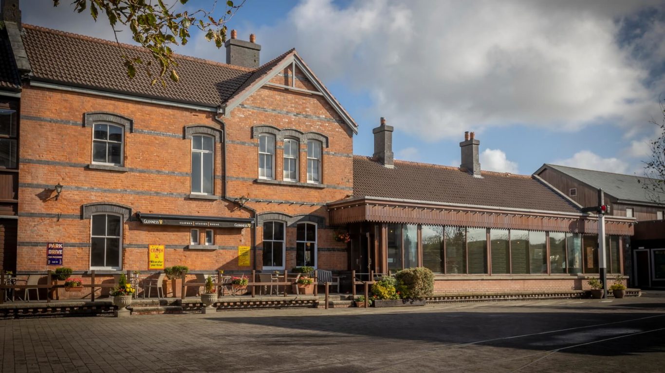 A two-story brick building with large windows and a glass extension, surrounded by potted plants, under a partly cloudy sky.