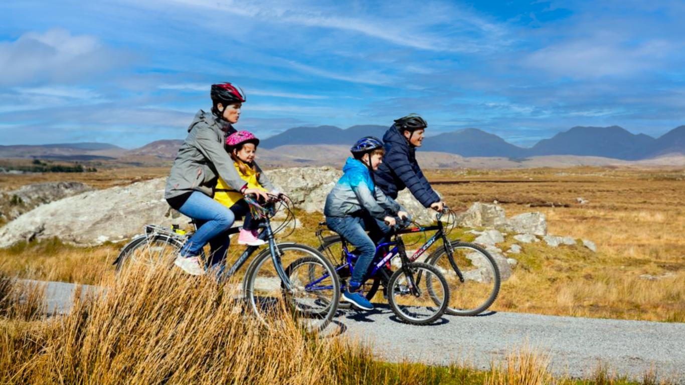 Four people, including two children, ride bicycles on a paved path through a grassy, open landscape with mountains in the background under a blue sky.