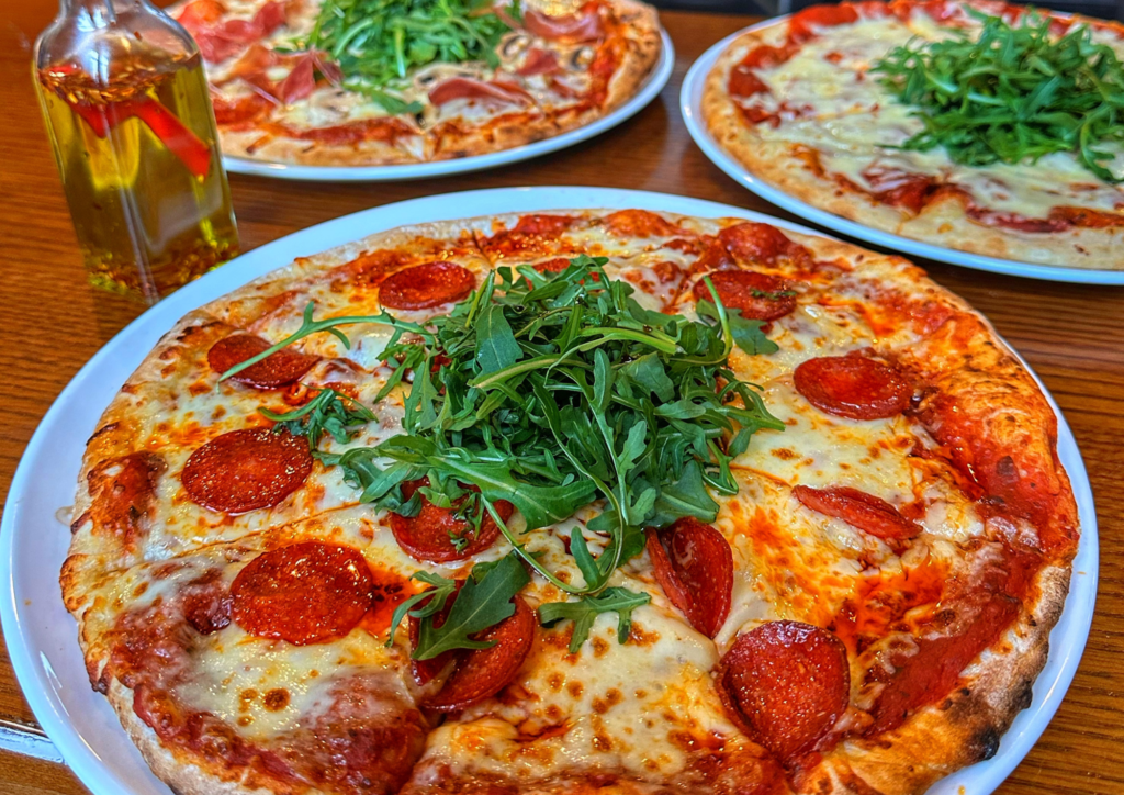 Three pizzas topped with arugula and various ingredients, including pepperoni, sit on white plates on a wooden table next to a bottle of olive oil with a red chili.