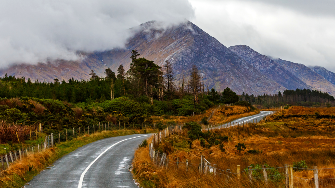 A winding road leads through grassy fields and past trees toward a tall, cloud-covered mountain under a gray sky.