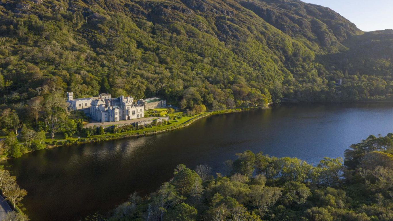 A large castle-like building sits beside a lake at the base of a forested hillside under clear skies.