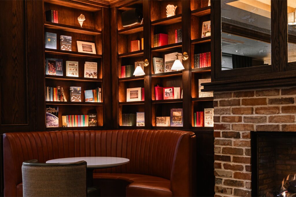A cozy corner with a round table and brown leather booth seating is set against wooden bookshelves filled with books and decor, next to a brick fireplace.