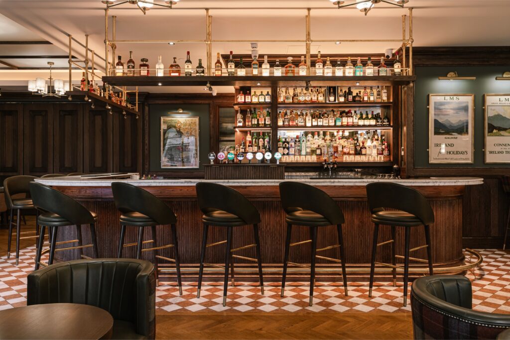 A stylish bar with five empty black chairs, a marble countertop, and shelves stocked with bottles and glasses behind the counter. The floor has a checkered pattern.