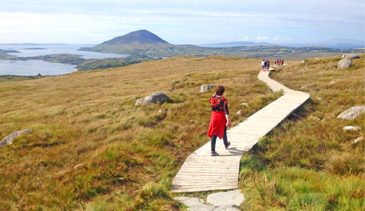 A person walks on a wooden boardwalk through grassy hills, with a group ahead and a mountain and water in the background under a partly cloudy sky.