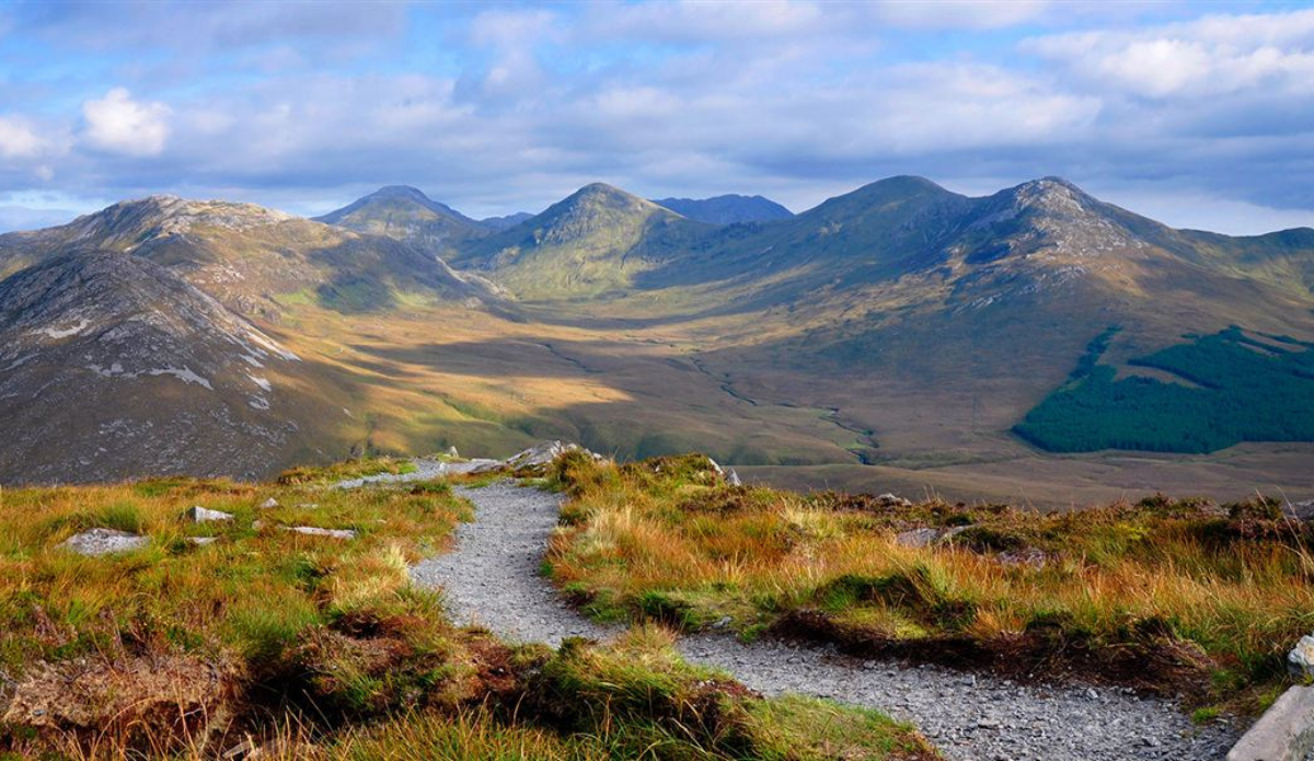 A winding gravel path leads through grassy hills toward a range of mountains under a partly cloudy sky.