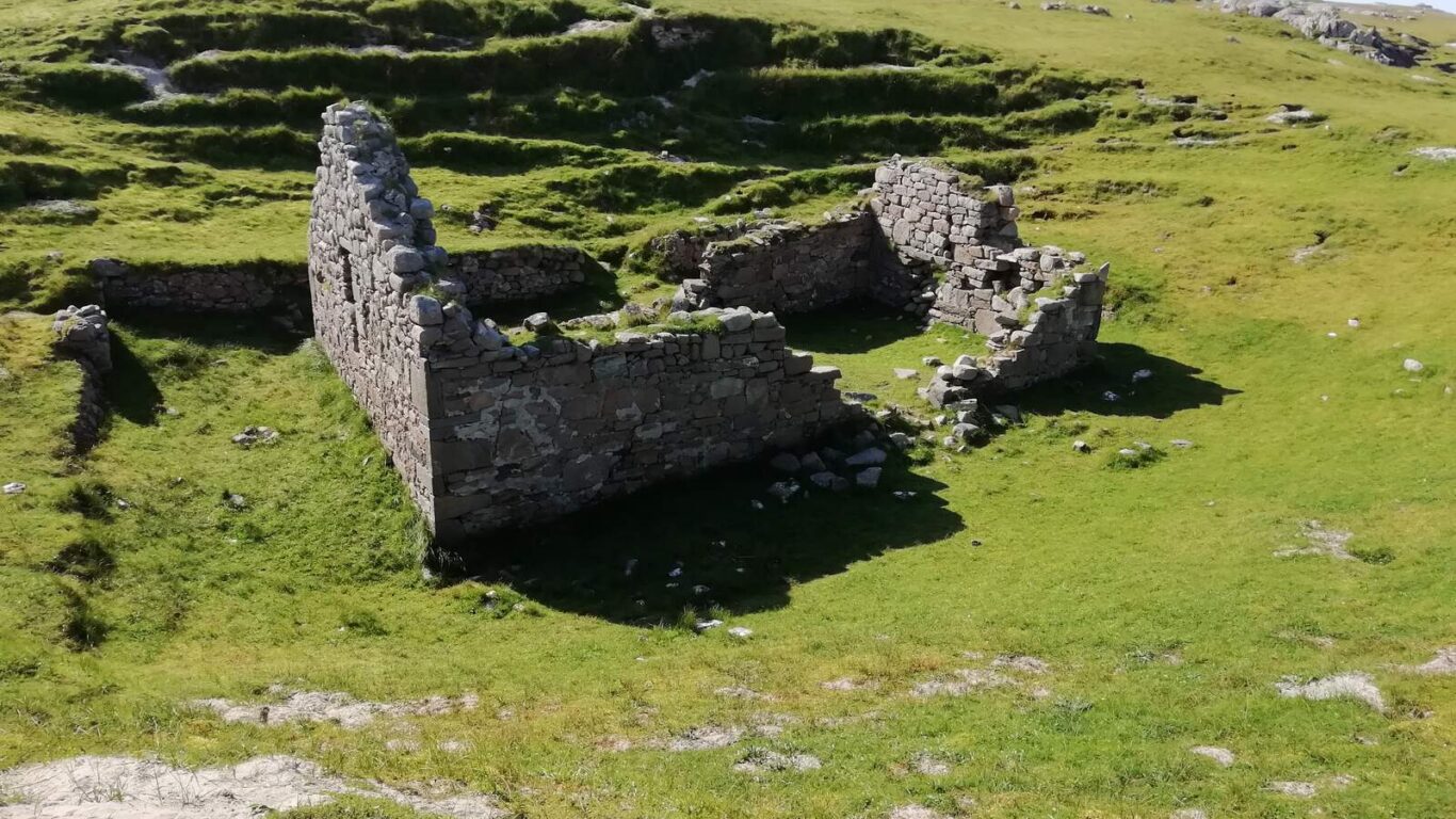 Stone ruins of a small building with partial walls stand on green grassy terrain under a clear sky.