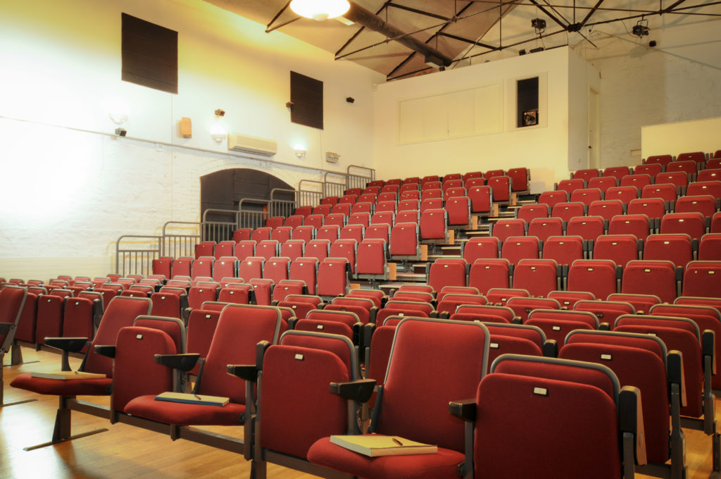 An empty lecture hall with rows of red seats, small desks attached, and a wooden floor, viewed from the front corner.