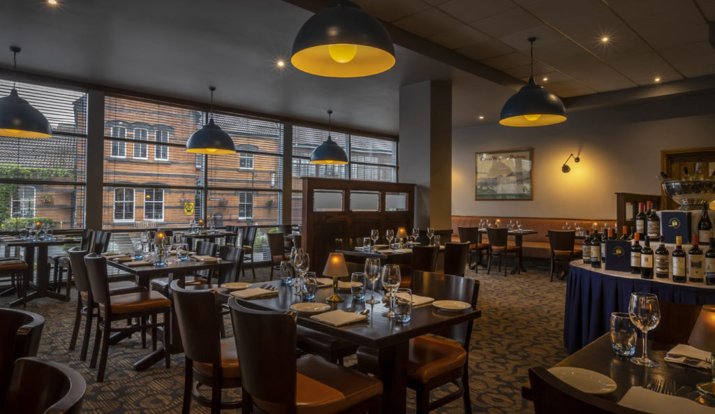 A restaurant dining area with neatly set tables, wooden chairs, large windows, pendant lights, and a table displaying bottles of wine.