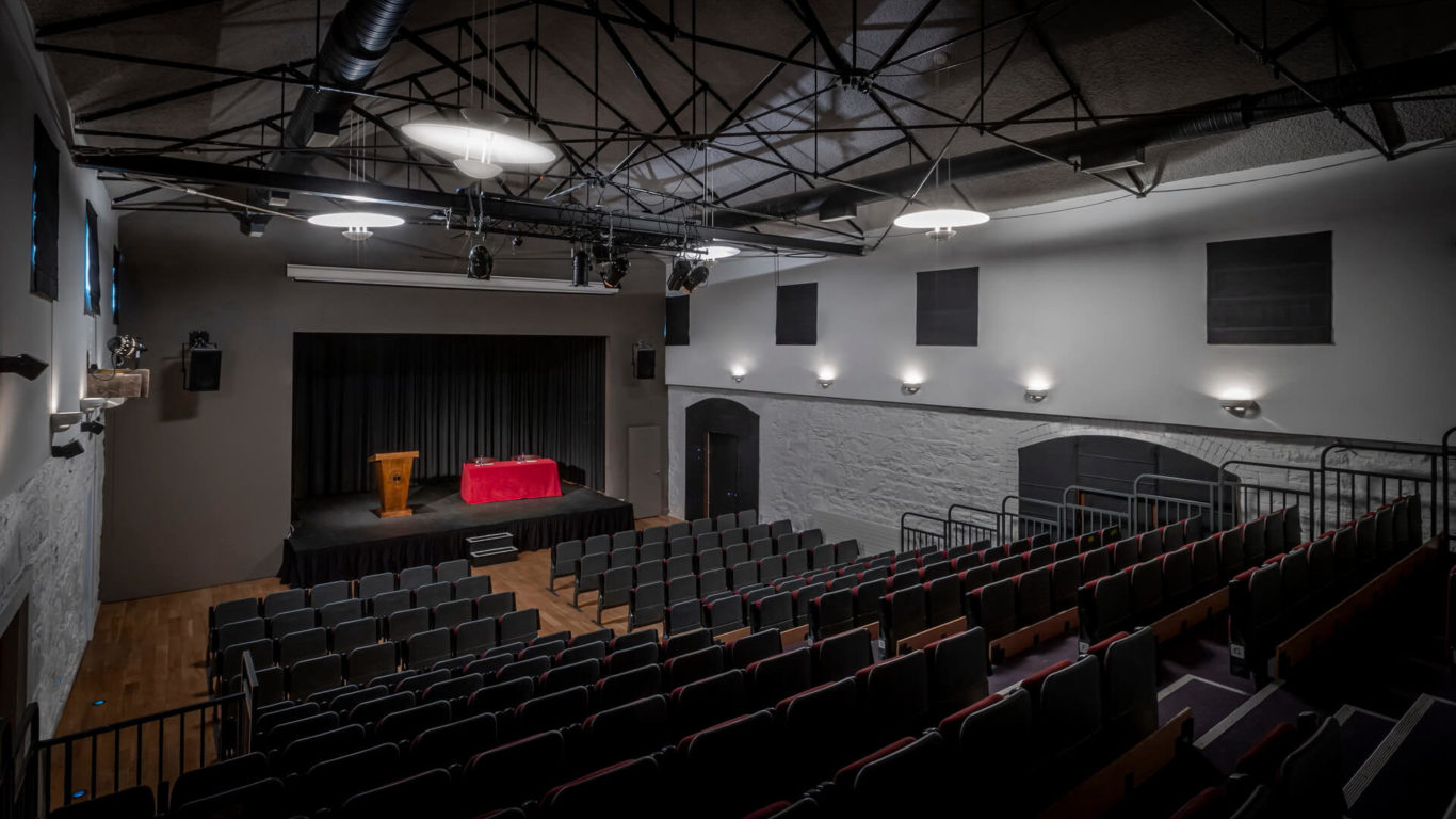 Empty auditorium with rows of seats facing a stage, which has a podium and a table covered with a red cloth under stage lighting.