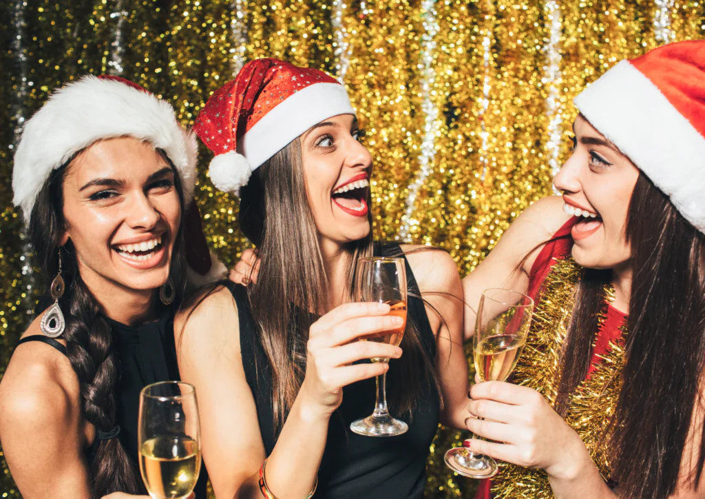 Jingle Night Celebration Three women wearing Santa hats hold champagne glasses and smile at each other, standing in front of a gold tinsel backdrop.