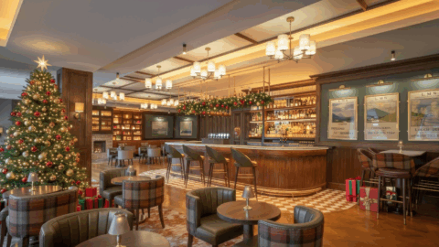 A cozy bar decorated for Christmas with a large tree, hanging garlands, wrapped gifts, and festive lights; empty tables and chairs are arranged around a central wooden bar.