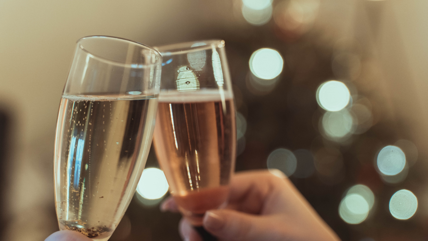 Two people clink glasses of sparkling wine during a cozy staycation, with a blurred background of festive lights.