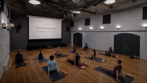A group of people practice yoga on mats in a spacious studio with wood floors, facing a screen that reads “Connemara Wellness Retreat.”.