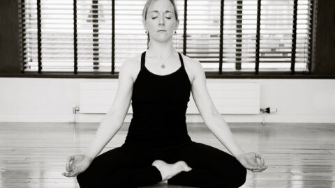 A woman sits cross-legged on a wooden floor, eyes closed, and hands resting on knees in a yoga meditation pose.