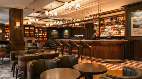 A warmly lit bar and lounge with wooden tables, leather chairs, high-backed barstools, and shelves stocked with bottles behind the bar.