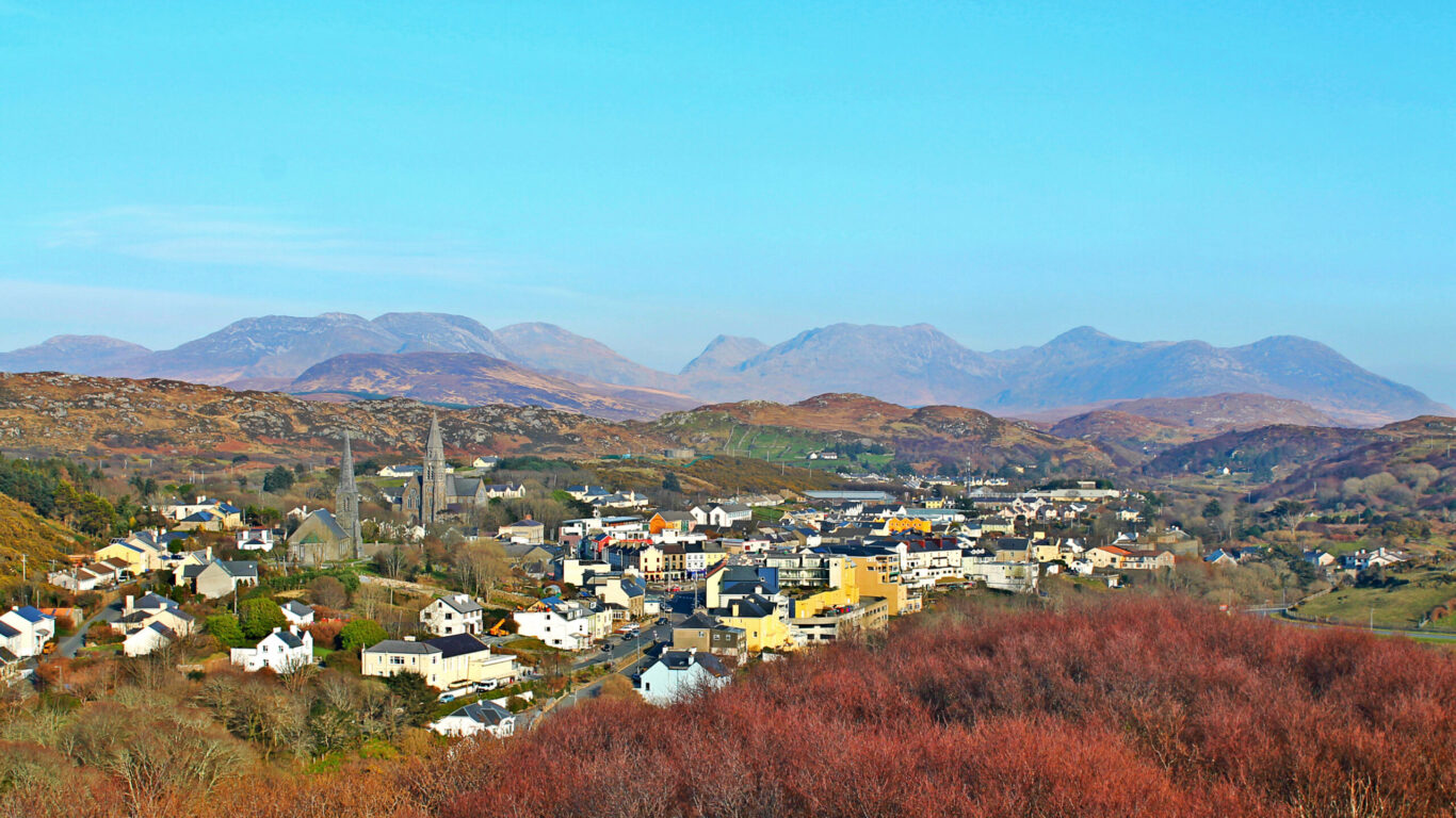 A small town with clusters of houses and buildings is set among rolling hills, with distant mountains under a clear blue sky.